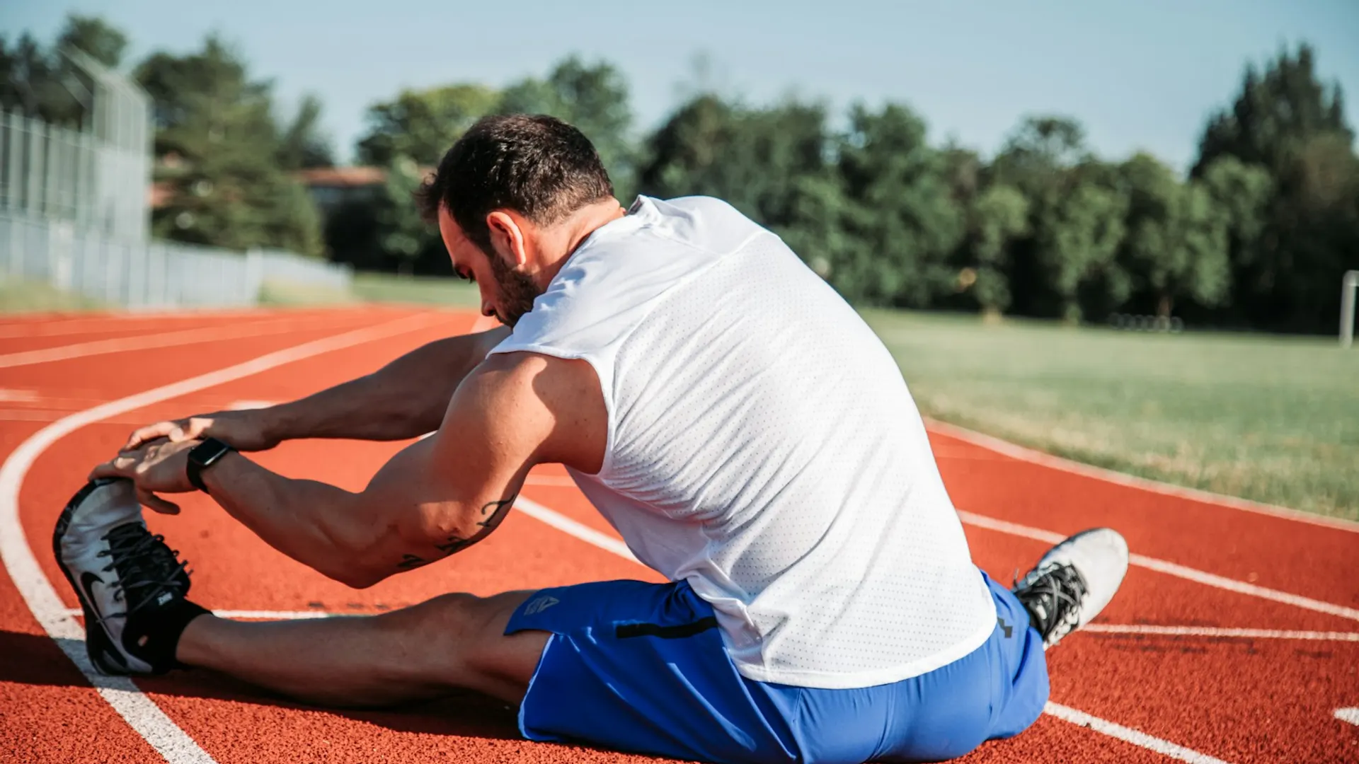 A man in a white tank top stretching toward his foot on a running track